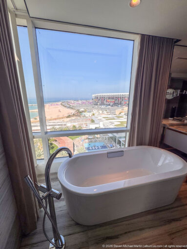 Freestanding white bathtub beside a floor-to-ceiling window, with a stadium and coastal development visible outside.
