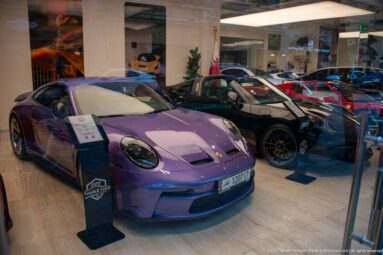 Purple Porsche 911 in a showroom with black and red sports cars displayed behind it.