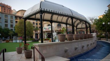 Covered stone fountain with black iron supports, white canopy, urn planters, and lion-head water spouts.