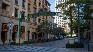 Green Plaza Andalucia gateway over a pedestrian street with restaurants, trees, balconies, and globe lamps.