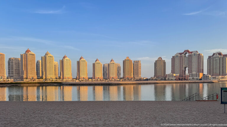Row of pale residential towers with red-roofed crowns reflected in calm water at The Pearl-Qatar.