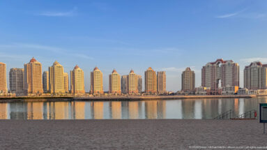 Row of pale residential towers with red-roofed crowns reflected in calm water at The Pearl-Qatar.