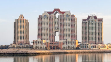 Three cream residential towers with red roofs and a large central arch beside the waterfront at The Pearl-Qatar.