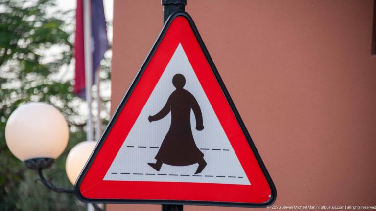 Red triangular pedestrian warning sign showing a figure in long traditional Gulf dress crossing road markings.