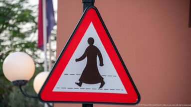 Red triangular pedestrian warning sign showing a figure in long traditional Gulf dress crossing road markings.