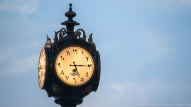 Ornate illuminated street clock with stylized Arabic numerals and dark decorative metalwork against a blue evening sky.