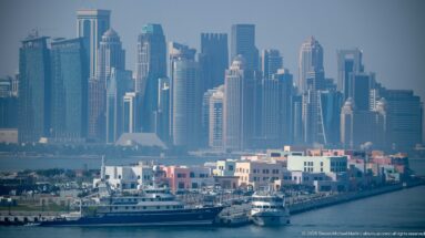 Dark luxury yacht and smaller boats beside pastel waterfront buildings with Doha’s West Bay towers behind.