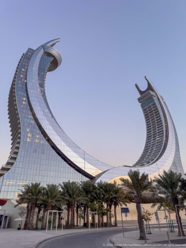 Low-angle view of Katara Towers with two sweeping curved glass façades rising above palm trees.