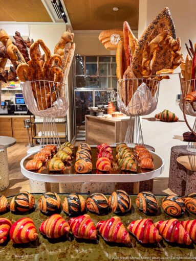 Hotel bakery buffet with baguettes, rustic breads, croissants, and rows of brightly striped pastries.