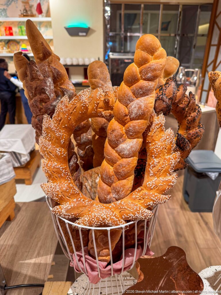 Basket filled with twisted rolls, sesame-topped flat breads, and rustic loaves at a hotel buffet.