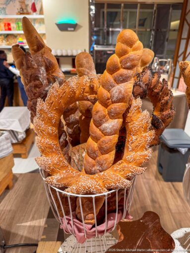 Basket filled with twisted rolls, sesame-topped flat breads, and rustic loaves at a hotel buffet.