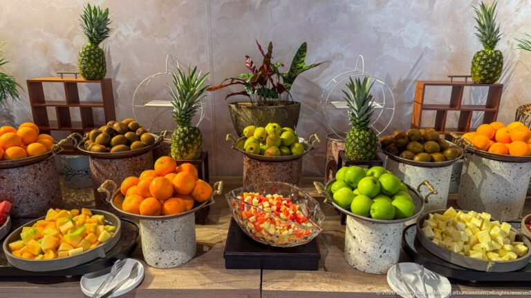 Hotel fruit buffet with pineapples, oranges, green apples, kiwifruit, and bowls of cut fruit in metal containers.