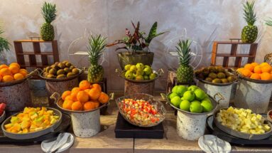 Hotel fruit buffet with pineapples, oranges, green apples, kiwifruit, and bowls of cut fruit in metal containers.