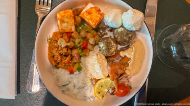 Overhead view of a buffet plate with hummus, fish, vegetables, grains, creamy salad, fried items, and lemon.