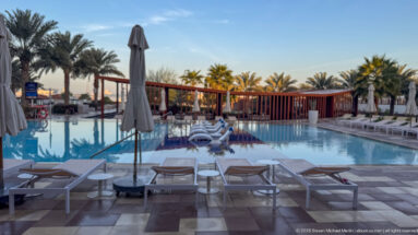 Wide resort swimming pool with cushioned loungers, closed umbrellas, palms, and white in-water chairs.