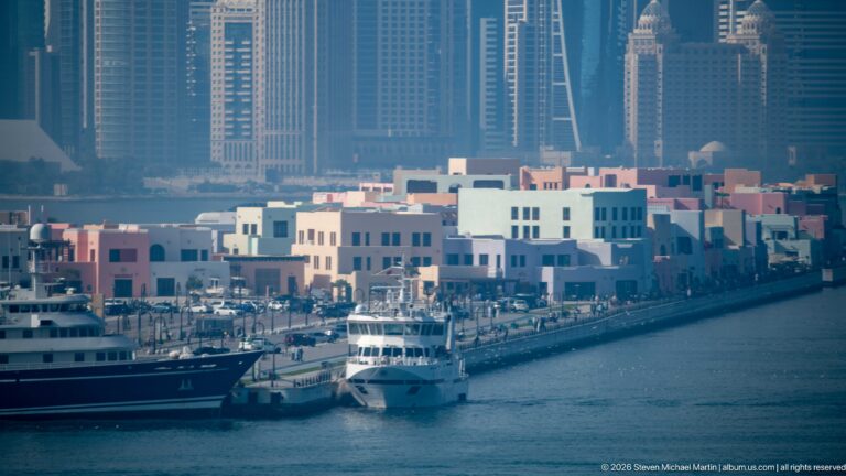 Pastel Mina District waterfront buildings and moored yachts with Doha’s high-rise skyline rising behind them.