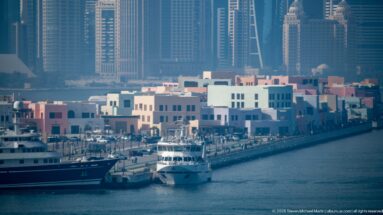 Pastel Mina District waterfront buildings and moored yachts with Doha’s high-rise skyline rising behind them.