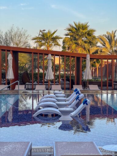 White in-water lounge chairs on a shallow resort pool ledge with closed umbrellas, palms, and a wood pergola.
