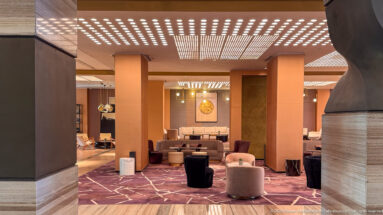 Warm airport lounge with rust-colored columns, cream seating, patterned carpet, dark tables, and rows of ceiling lights.