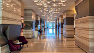 Long hotel corridor with striped stone walls, burgundy chairs, planters, and many suspended geometric ceiling lights.