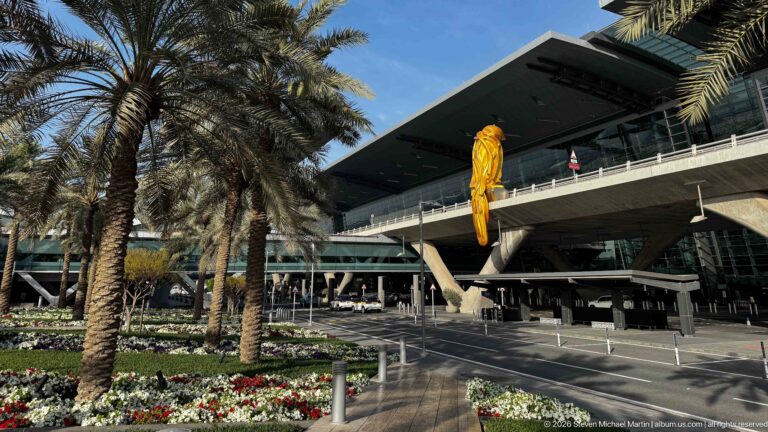 Airport terminal canopy above a curbside road, framed by date palms, flower beds, and a large yellow hanging emblem.