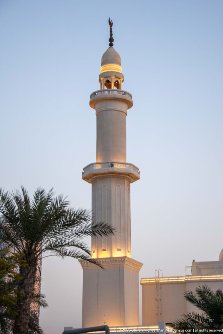 White cylindrical minaret with stacked balconies and a warmly lit crown rises behind palm fronds at dusk.