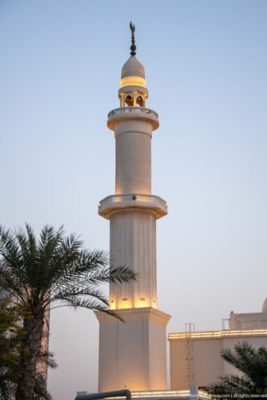 White cylindrical minaret with stacked balconies and a warmly lit crown rises behind palm fronds at dusk.