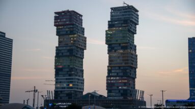 Two tall stacked-box towers rise side by side at dusk above cranes and lower buildings.