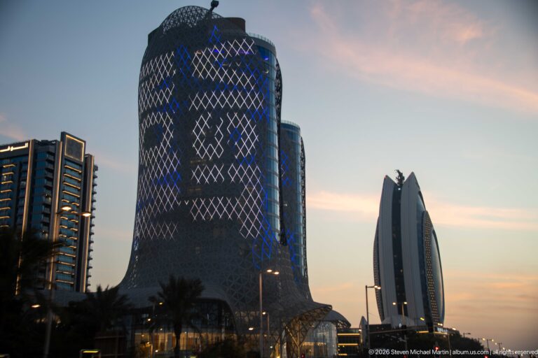 Rounded tower with blue diamond-lattice lighting stands beside a slender sculptural high-rise at dusk.