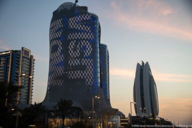 Rounded tower with blue diamond-lattice lighting stands beside a slender sculptural high-rise at dusk.