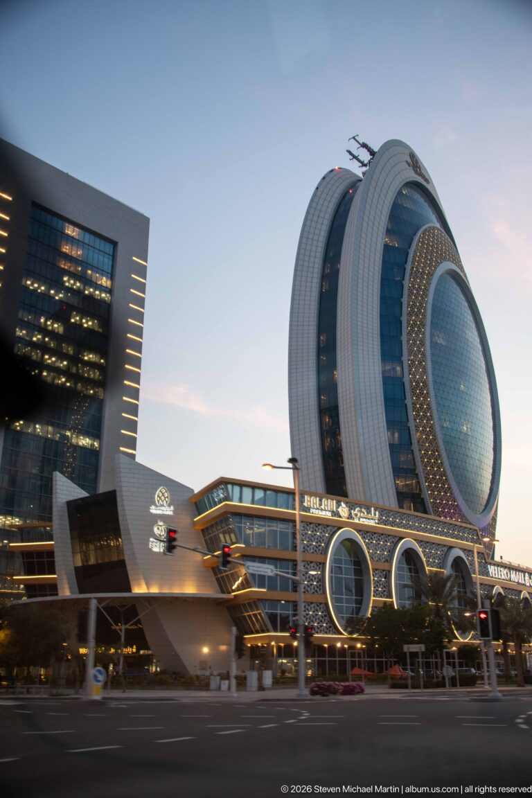 Large oval glass hotel tower with white ribs and perforated elliptical screen rises above an illuminated podium.