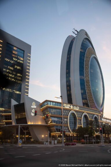 Large oval glass hotel tower with white ribs and perforated elliptical screen rises above an illuminated podium.