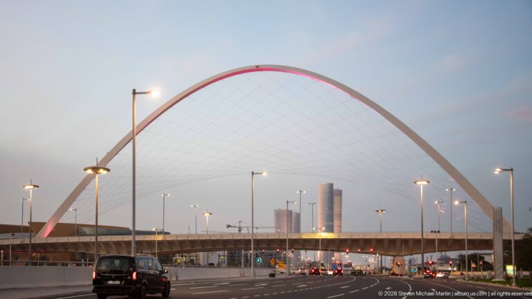 Large white cable-supported arch spans a multi-lane road with distant towers visible beyond.