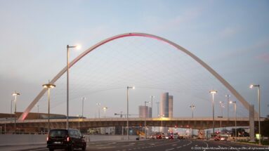 Large white cable-supported arch spans a multi-lane road with distant towers visible beyond.