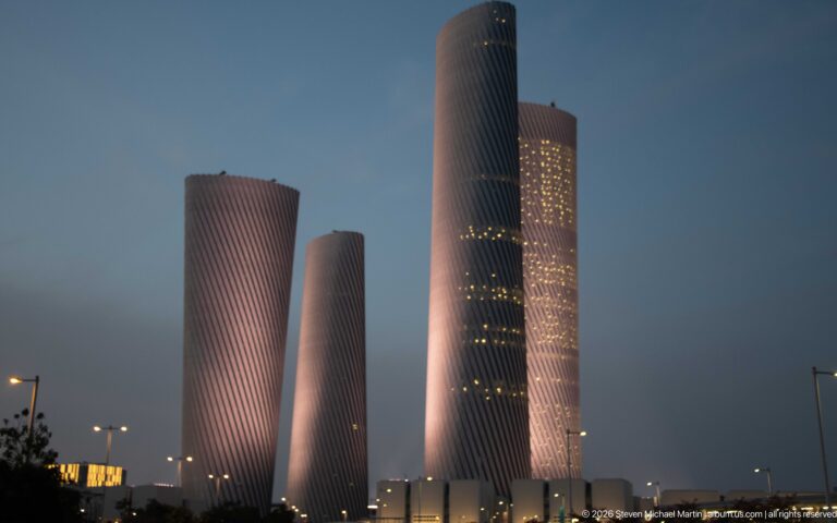 Four softly lit twisting Lusail Plaza Towers rise above low buildings at twilight.