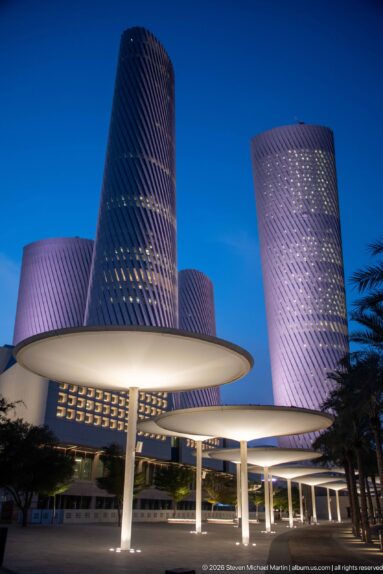 White circular canopy roofs and columns frame purple-lit Lusail Plaza Towers at blue hour.