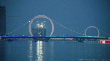 Illuminated bridge spans the water beneath two circular cable arches, framing a tower in the larger ring.