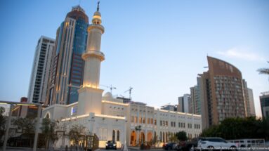 Illuminated mosque with tall minaret and arched white façade stands among modern high-rise buildings at dusk.