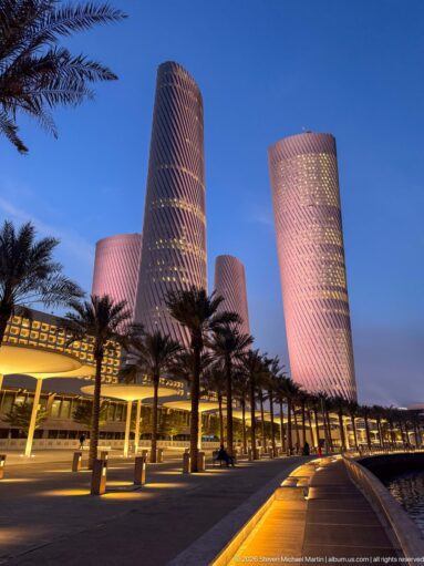 Wide blue-hour view of pink-lit Lusail Plaza Towers behind a palm-lined promenade.