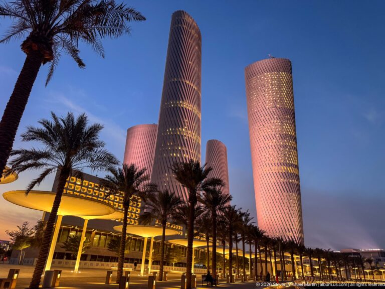 Pink and lavender Lusail Plaza Towers rise above palms and warmly lit podiums at blue hour.