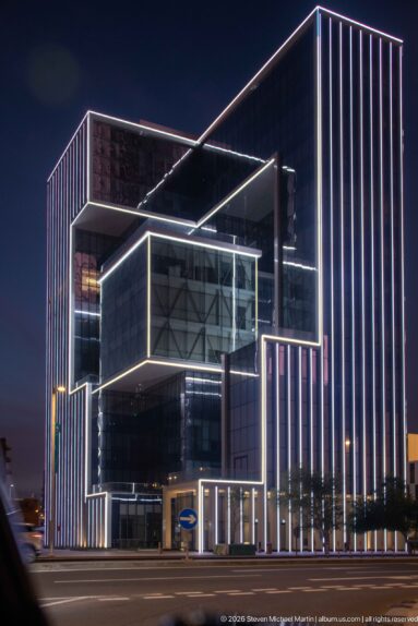 Dark glass office building with stacked volumes outlined by continuous white LED lighting at night.
