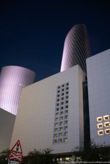 Twisting Lusail towers rise behind white podium walls with vertical and square window patterns.
