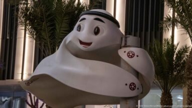 Smiling white mascot shaped like a Gulf head covering stands beside palms at night.