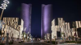 Two violet-lit twisting Lusail Plaza Towers rise above a broad boulevard lined with modern buildings.