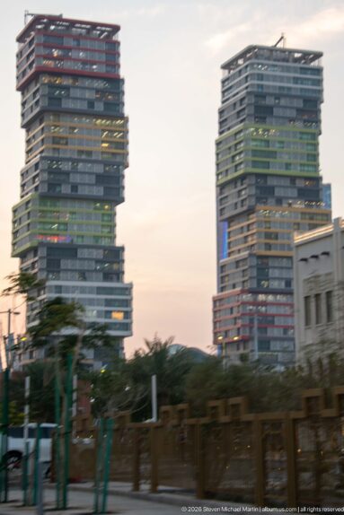 Two modern stepped high-rises with projecting horizontal floor plates rise behind palms at dusk.