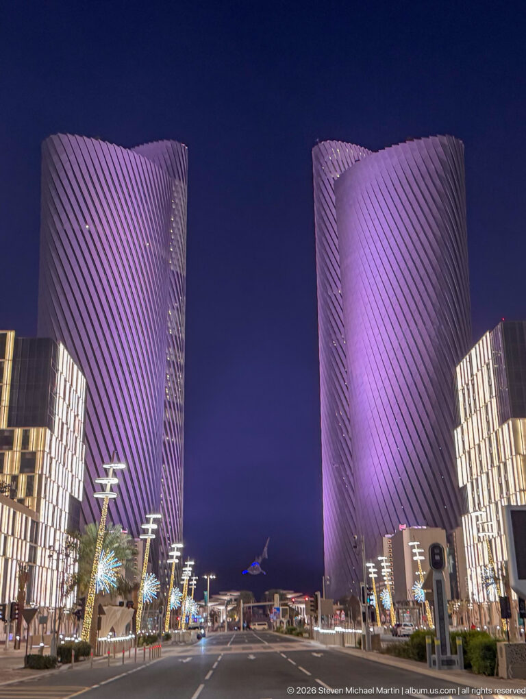 Purple-lit Lusail Plaza Towers frame a boulevard lined with modern low-rise buildings and streetlights.