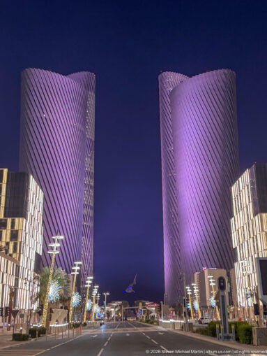 Purple-lit Lusail Plaza Towers frame a boulevard lined with modern low-rise buildings and streetlights.