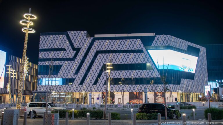 Angular modern complex with diamond-patterned façades rises above illuminated storefronts and traffic at night.