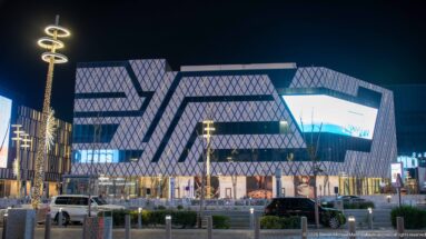 Angular modern complex with diamond-patterned façades rises above illuminated storefronts and traffic at night.