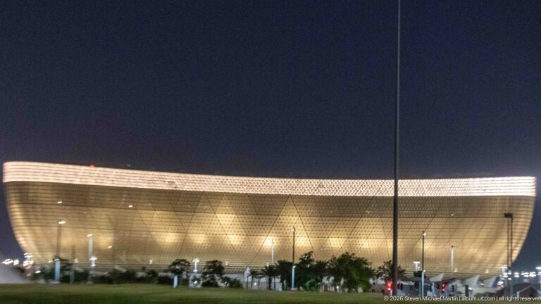 Golden bowl-shaped Lusail Stadium glows at night behind palms and roadway lights.